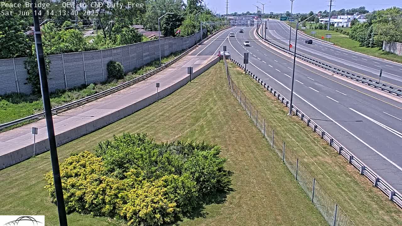 A highway scene on a sunny day shows several cars driving on a multi-lane roadway that curves gently to the left, bordered by grassy areas and a chain-link fence.