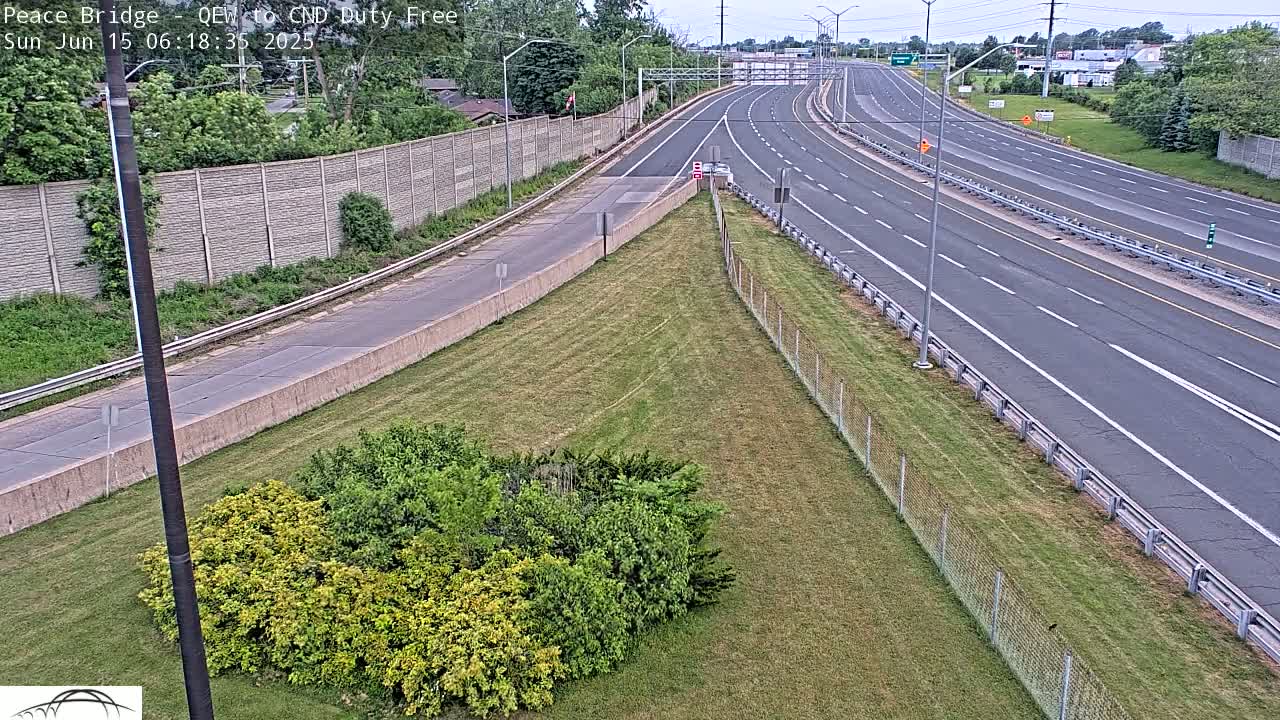 A highway with several lanes of traffic stretches into the distance under a partly cloudy sky, bordered by grassy areas and a chain-link fence.