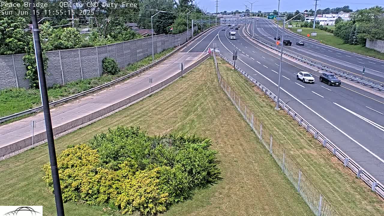 A high-angle view shows a highway with several vehicles traveling on a sunny day, alongside a grassy area bordered by a chain-link fence and shrubs.