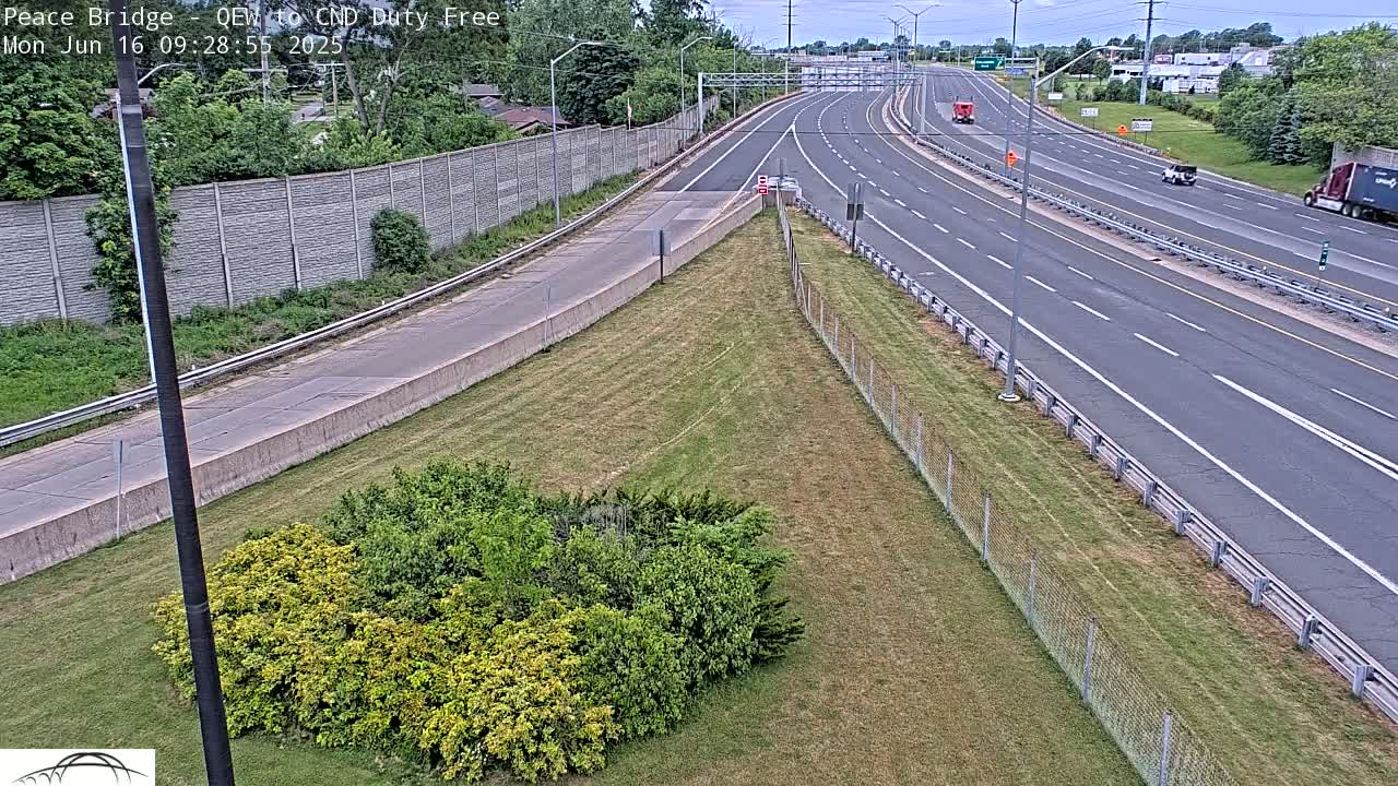 A multi-lane highway with sparse traffic on a partly cloudy day is bordered by a grassy area with shrubs and a chain-link fence.