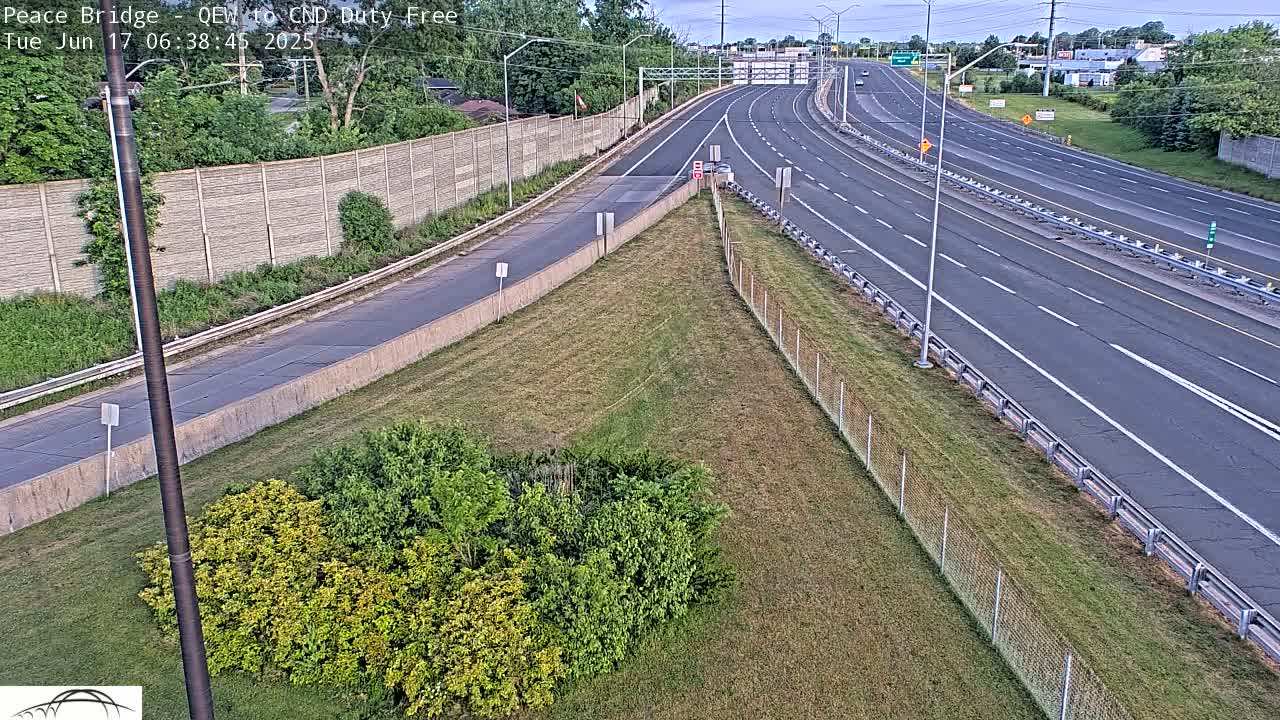 An aerial view of a highway with a few cars, under a partly cloudy sky, shows a grassy area with shrubs next to a concrete barrier.