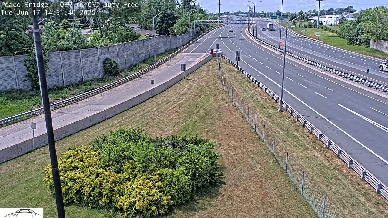 A highway with sparse traffic on a partly sunny day, bordered by a grassy area with shrubs and a chain-link fence.