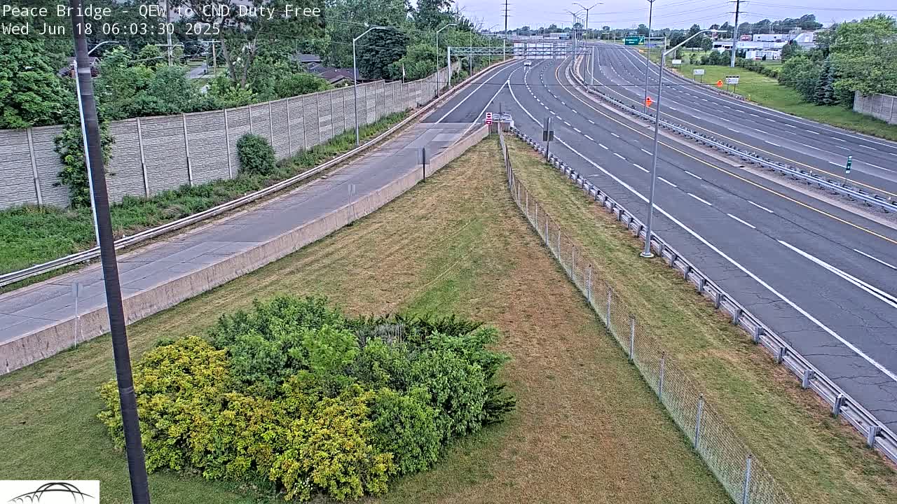 A high-angle view of a multi-lane highway on a partly sunny day, with grassy areas and a chain-link fence separating the road from a patch of shrubs.