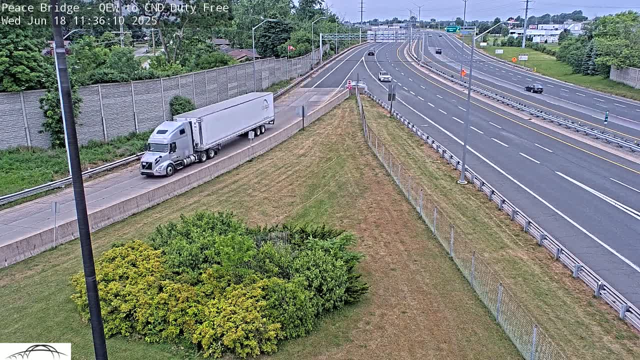 A semi-truck travels on a multi-lane highway on a partly sunny day, with grassy areas and shrubs visible beside the road.