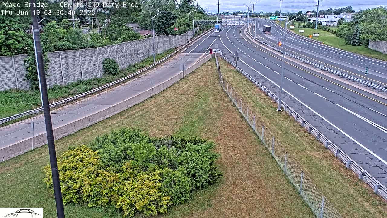 A mostly empty highway with a few vehicles visible stretches into the distance under a partly cloudy sky, bordered by grassy areas and chain link fencing.