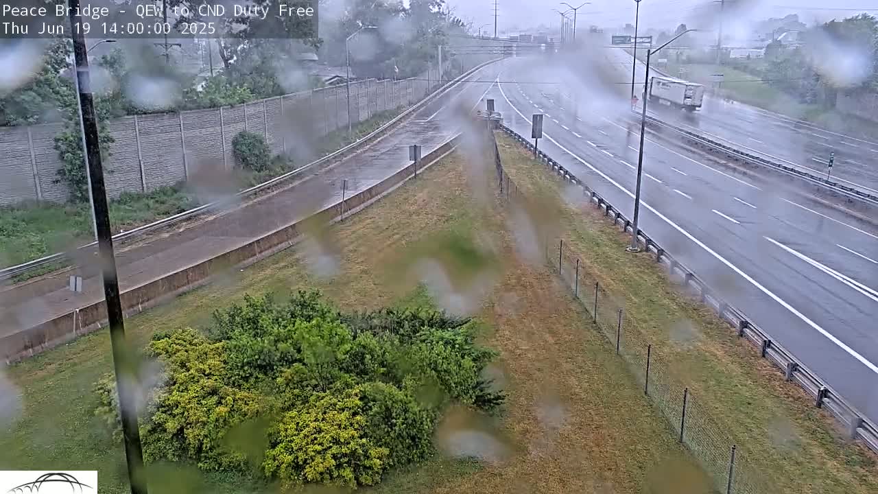 A rain-slicked highway merges with an off-ramp, bordered by grassy areas and a chain-link fence, with sparse traffic visible under an overcast sky.
