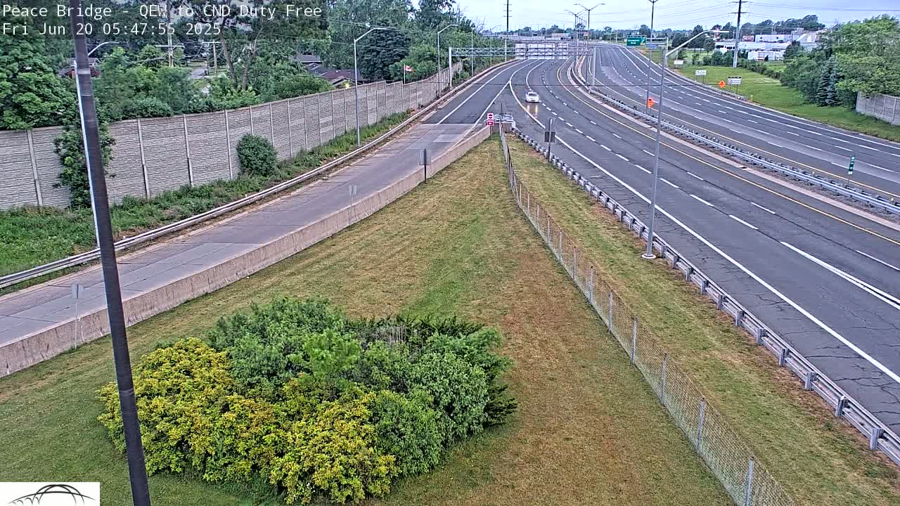 A highway with a few vehicles is seen from an elevated vantage point on a partly sunny day, with a grassy area and chain link fence separating the highway and viewer.