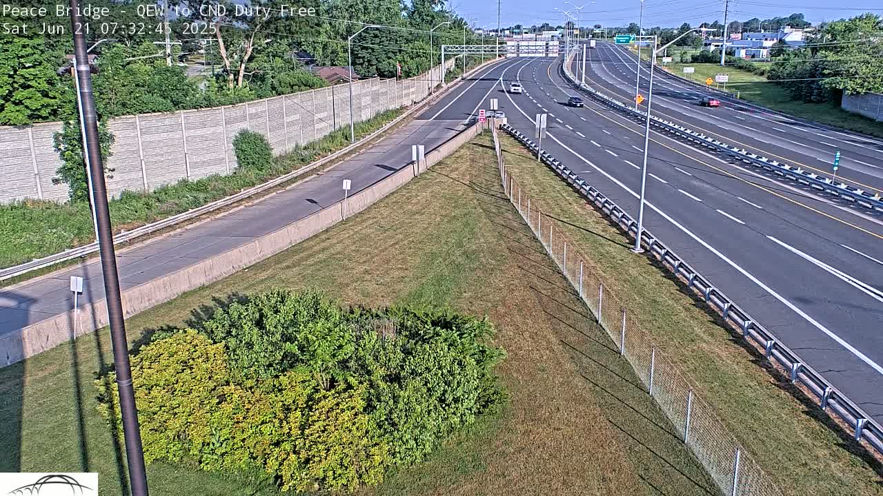 A multi-lane highway with sparse traffic on a sunny day, bordered by a grassy area with shrubs and a chain-link fence.