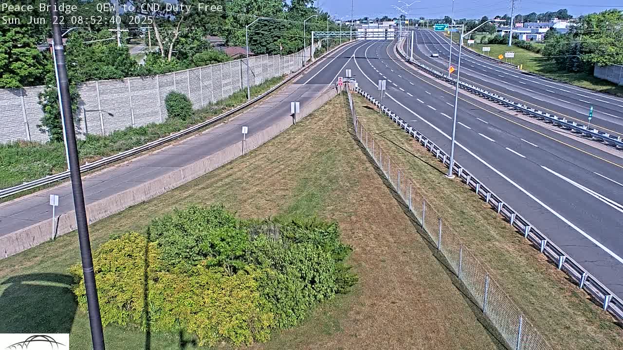 A multi-lane highway stretches into the distance under a bright, sunny sky, bordered by a grassy median and chain-link fences.
