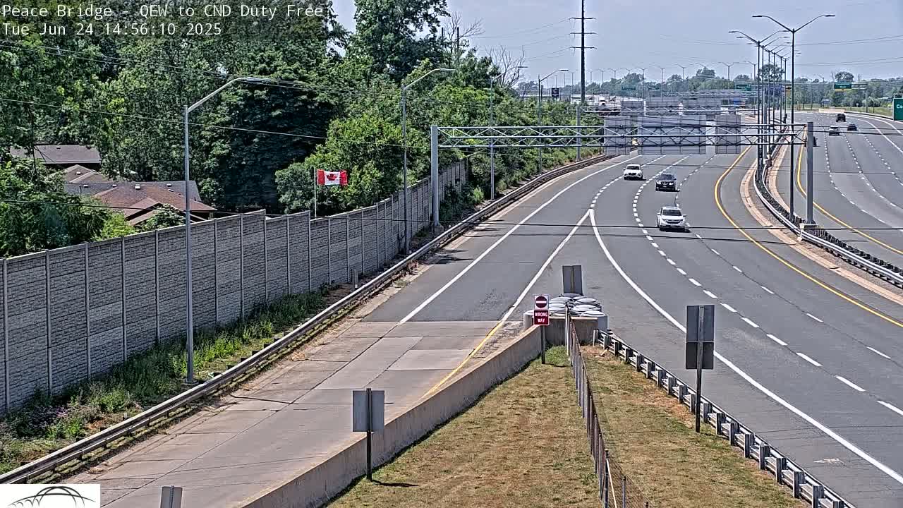 A multi-lane highway with several cars driving on a sunny day, bordered by a tall concrete wall and lush green vegetation.