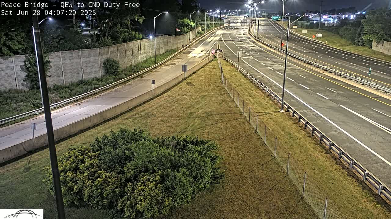 A nighttime view of a multi-lane highway merging onto another highway, surrounded by grassy areas and a chain link fence.