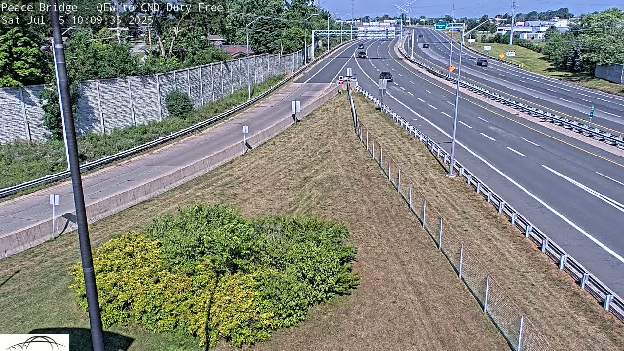 A highway with several cars driving on a sunny day, viewed from an elevated vantage point showing a grassy area and chain link fence beside the road.