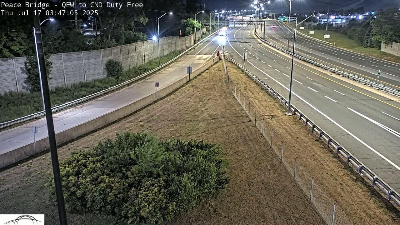 A nighttime high-angle view shows a multi-lane highway with sparse traffic, bordered by a chain-link fence and dry, brown grass.