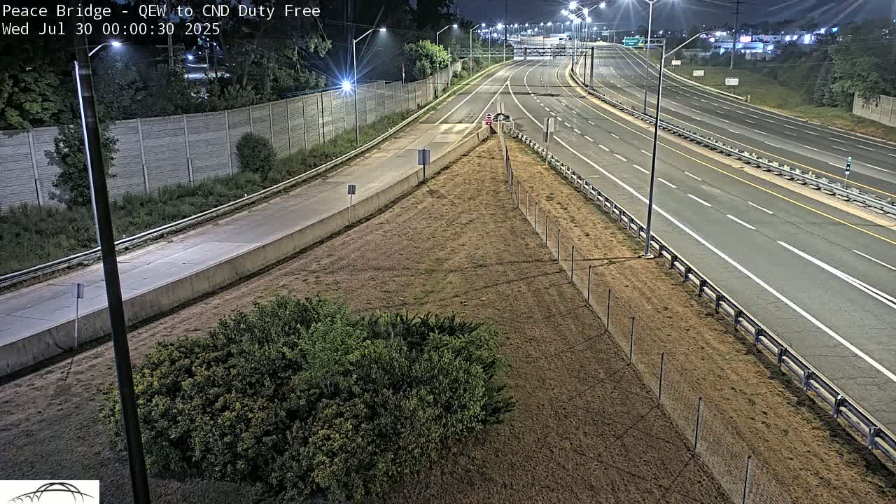 A nighttime high-angle view shows a multi-lane highway with sparse traffic, bordered by a chain-link fence and dry, brown grass.
