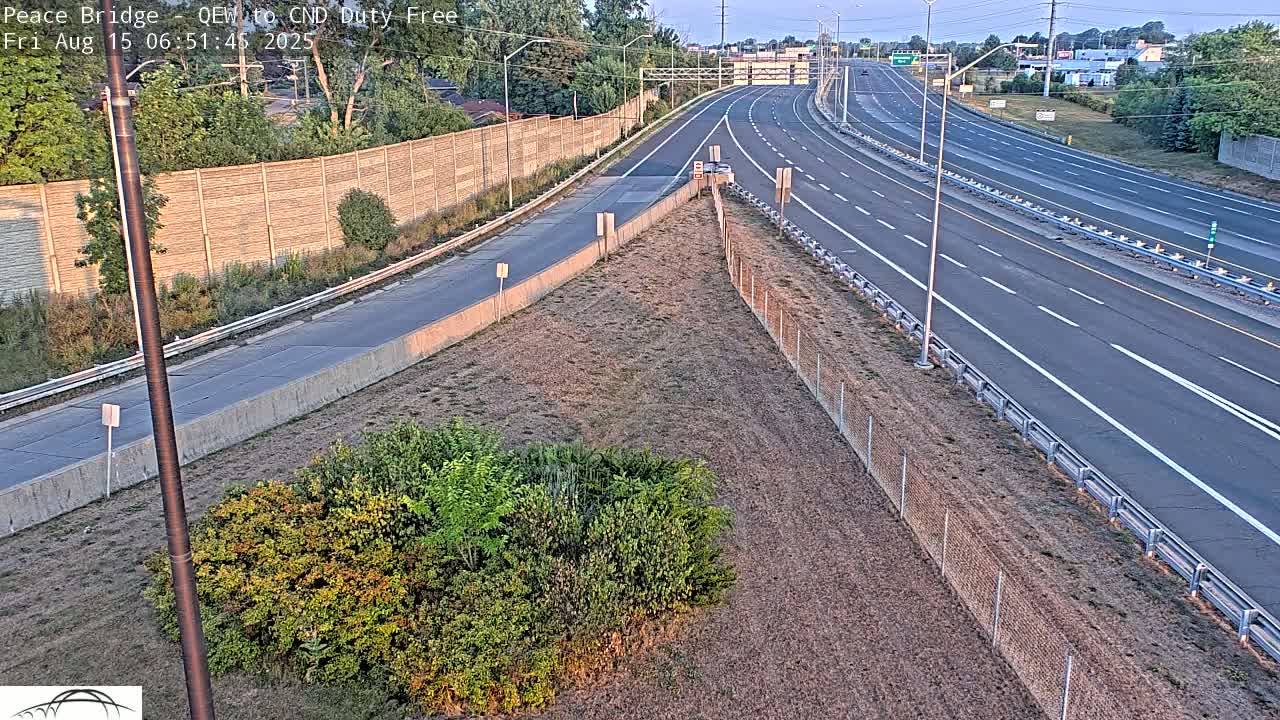 A nighttime high-angle view shows a multi-lane highway with sparse traffic, bordered by a chain-link fence and dry, brown grass.
