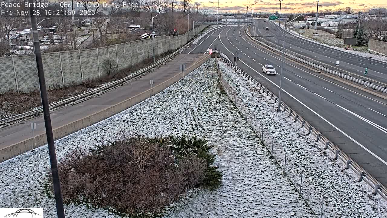 A multi-lane highway, a smaller road, and railway tracks are visible from an elevated perspective, with a thin, patchy layer of snow on the ground under a partly cloudy sky with hints of orange near the horizon.