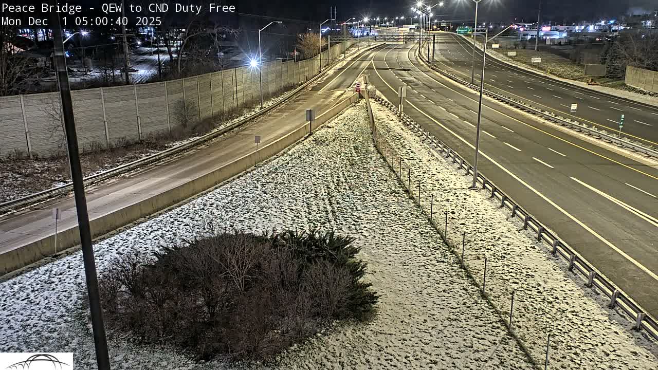 A nighttime view reveals a multilane highway approach lightly dusted with snow on the ground and surrounding areas, illuminated by numerous streetlights, with bare trees and fences visible along the sides.