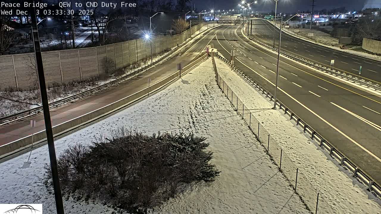 A multi-lane highway at night is shown with snow covering the ground and barriers beside the clear driving lanes, illuminated by numerous streetlights under a dark sky.