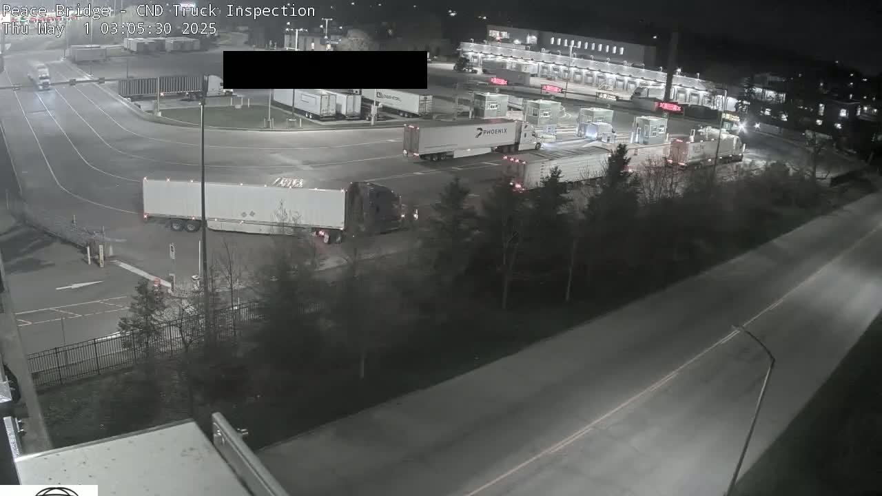 At night, several semi-trailer trucks are parked and moving in a large, well-lit area near a road.