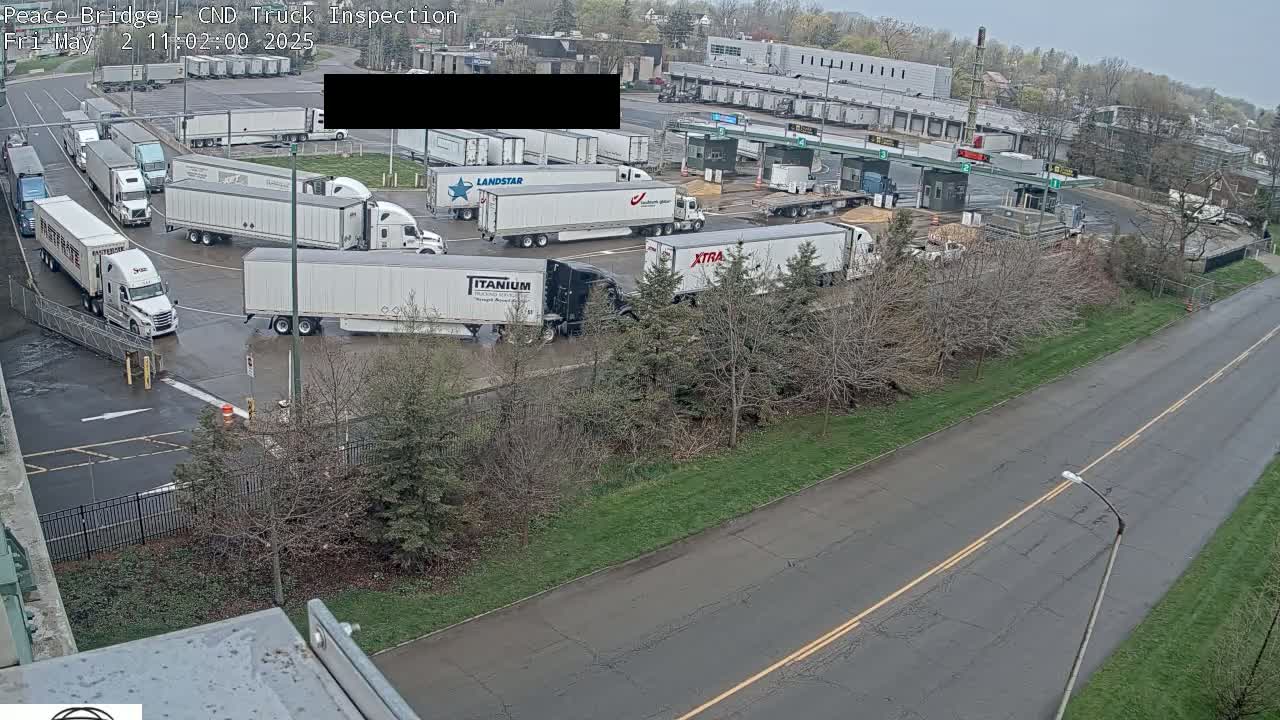 A wet parking lot is filled with numerous semi-trailer trucks near a border crossing, under an overcast sky.