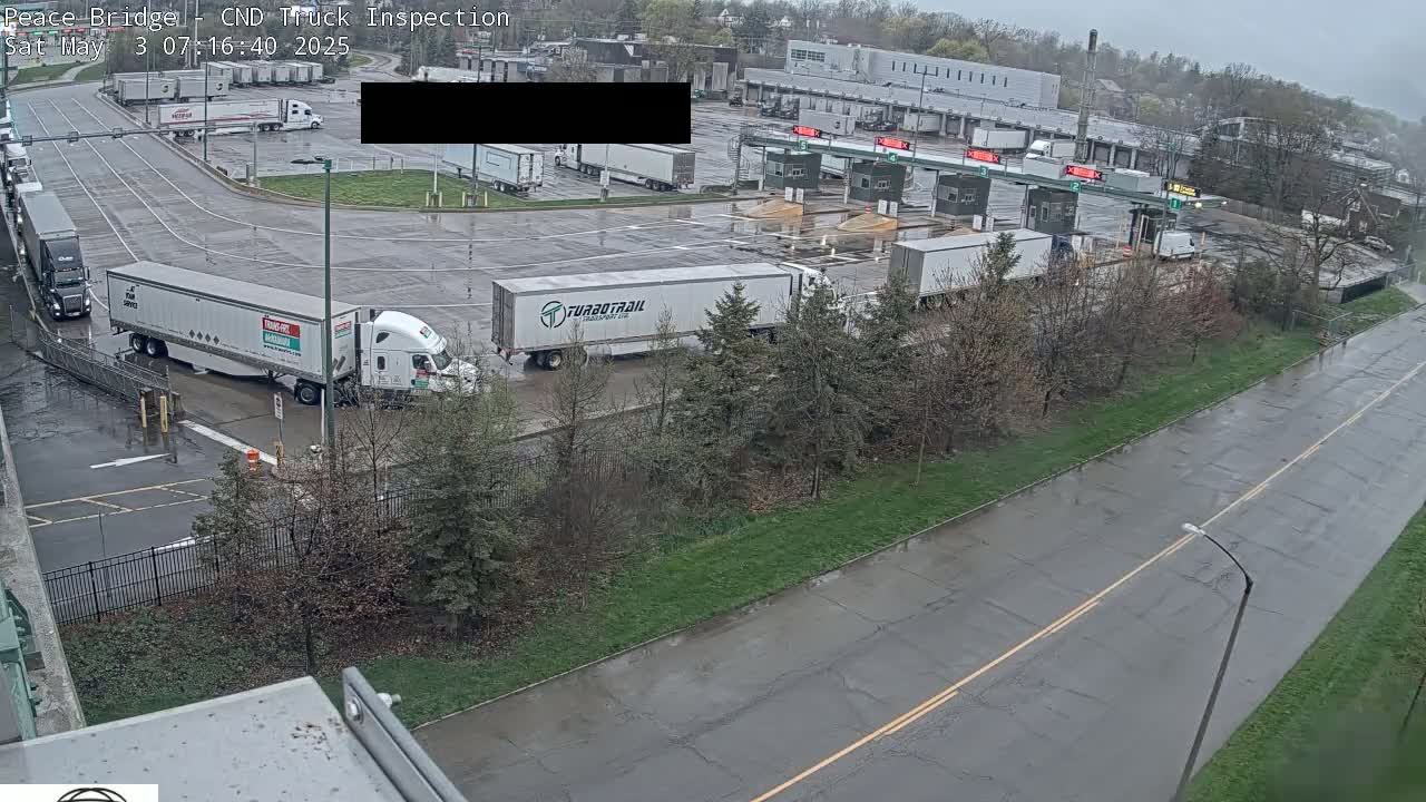 A wet, overcast day at a truck inspection station shows several semi-trucks waiting in a parking lot and at inspection booths.