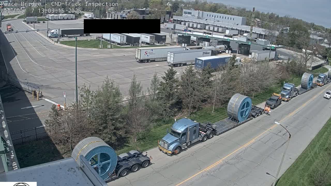 Several large trucks carrying spools of cable are on a road next to a truck inspection facility on a partly sunny day.