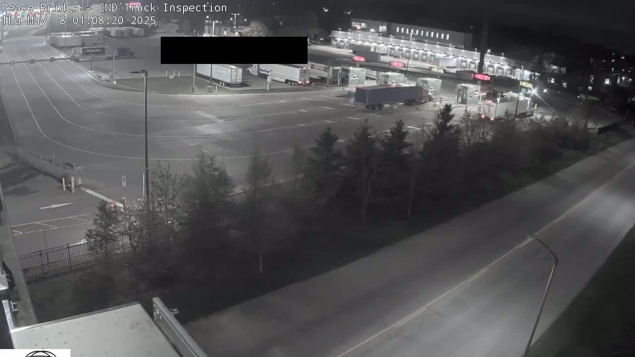 A nighttime aerial view shows a truck inspection area with several semi-trucks, a roadway, and surrounding landscaping.