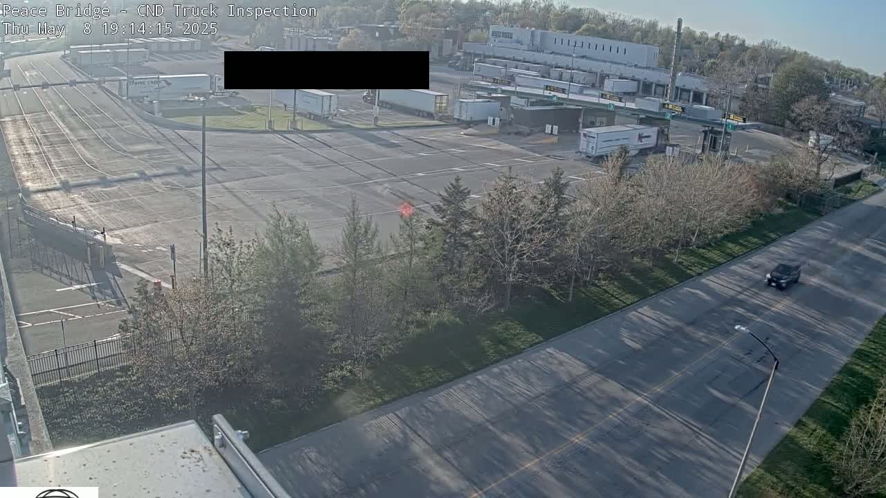 A truck inspection station with numerous semi-trucks parked in a large lot on a sunny day, viewed from above, with a road and trees in the foreground.