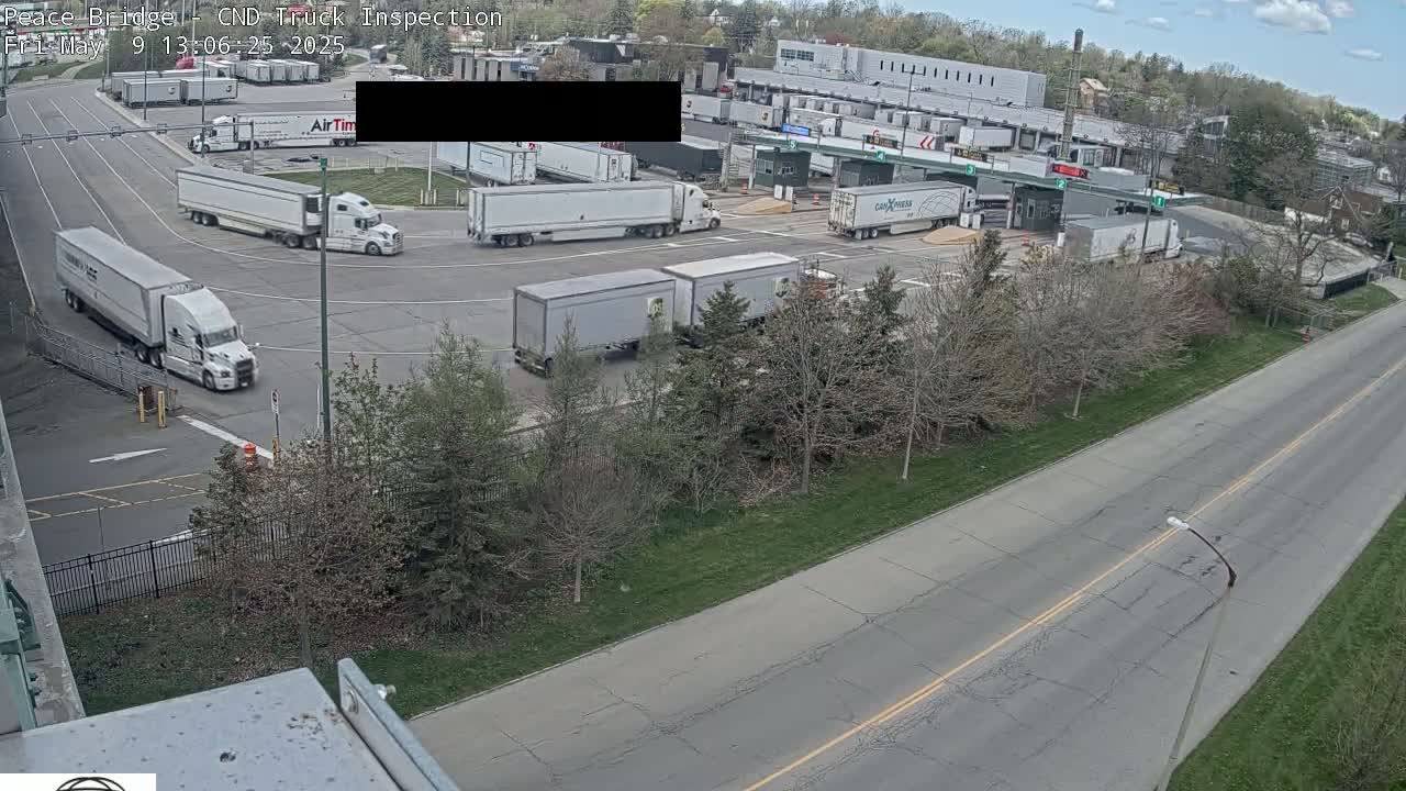 Several semi-trucks are parked and moving in a paved area near a border crossing on a partly cloudy day.