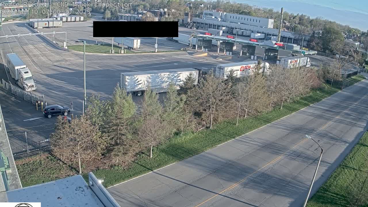 A truck inspection station with several semi-trucks and a dark-colored car visible, on a sunny day with many bare trees lining a roadway.