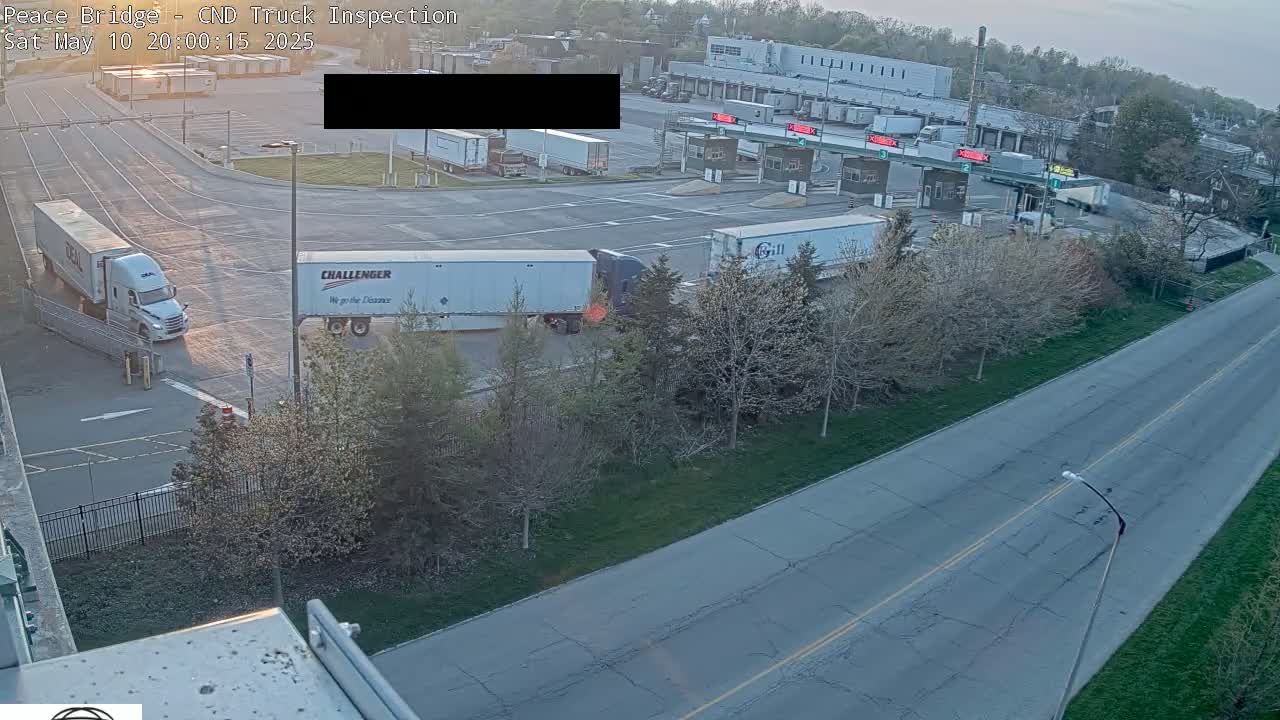 Several semi-trailer trucks are at a border crossing inspection area at sunrise, with some trucks waiting in line and others undergoing inspection.