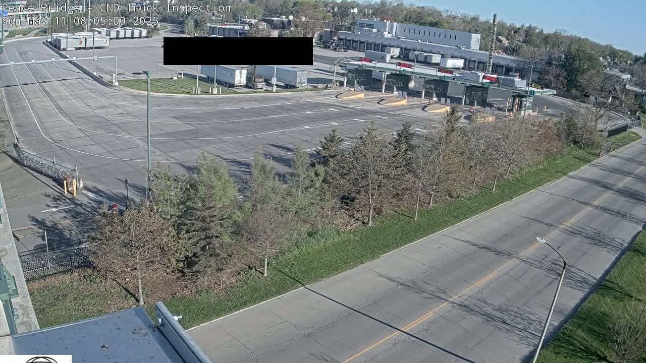 An overhead view shows a truck inspection station with several trucks parked, a roadway leading to it, and a row of trees alongside a road.