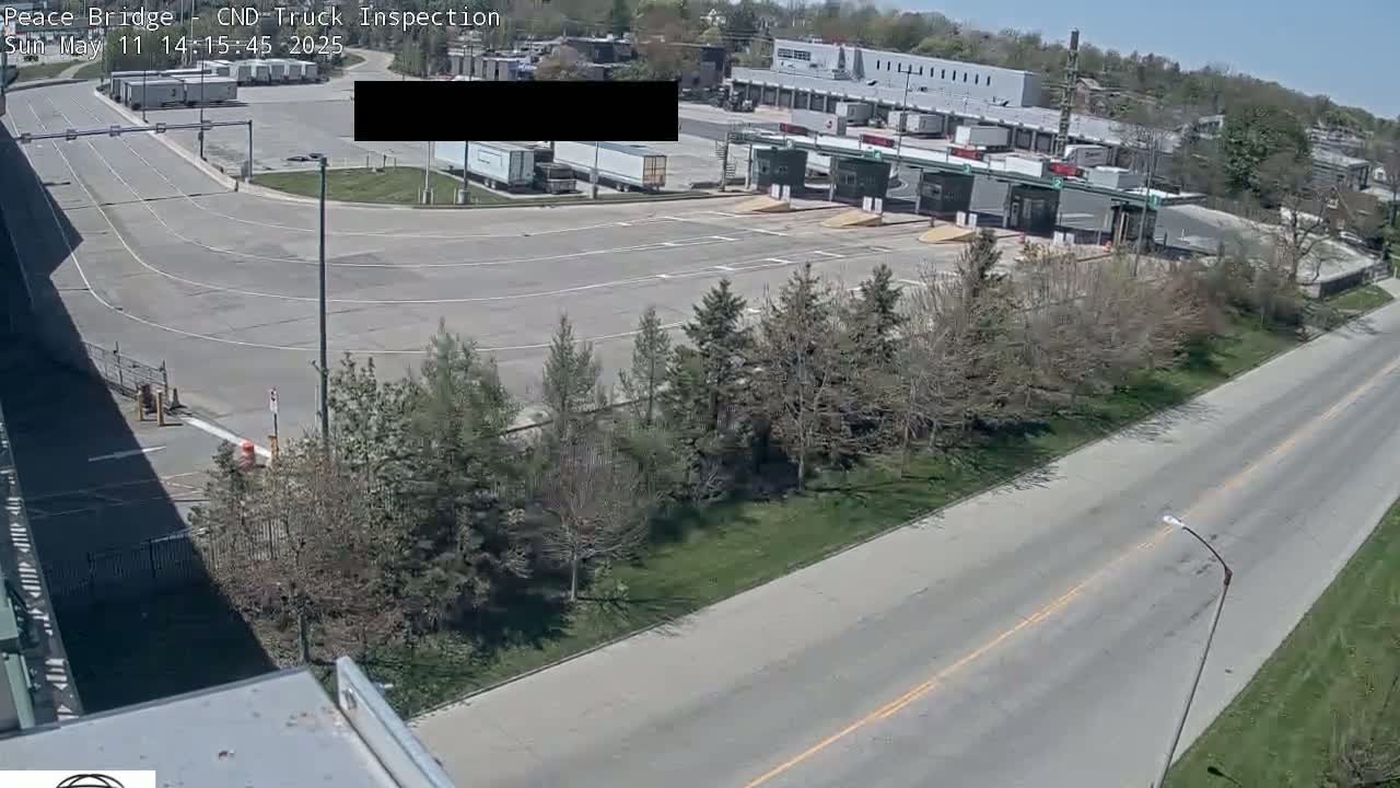A high-angle view shows a mostly empty truck inspection area on a sunny day, with several evergreen trees lining a road in the foreground.