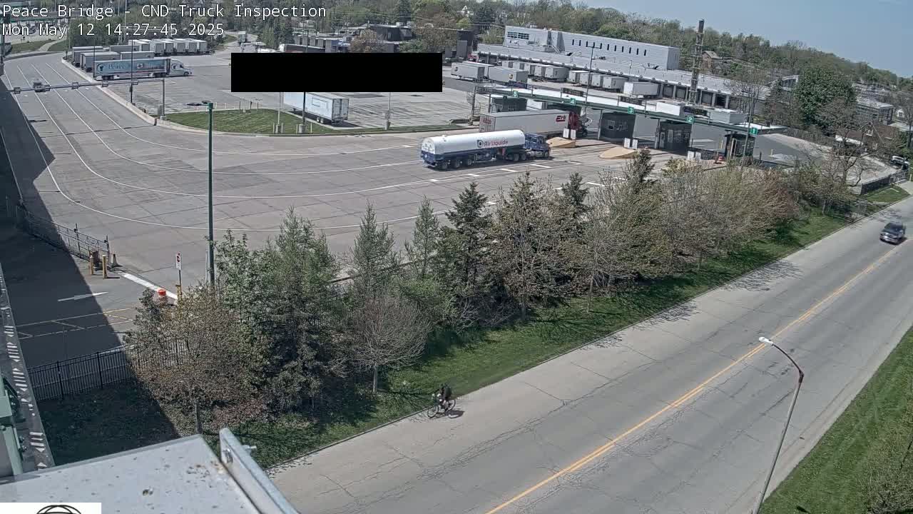 A high-angle view shows a truck inspection station on a sunny day, with several trucks and a tanker in the lot, and a cyclist riding along a road near a grassy area.