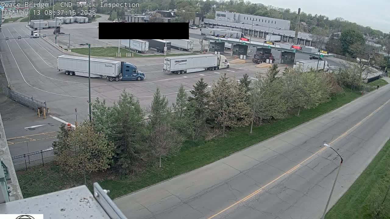 Several semi-trucks are parked and moving in a truck inspection area, under a partly cloudy sky.