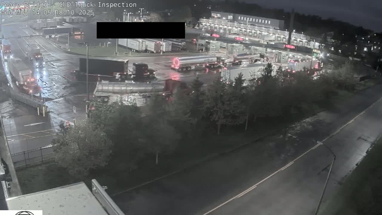 A nighttime, wet scene shows numerous semi-trucks at a truck inspection station, with some trucks moving and others parked.