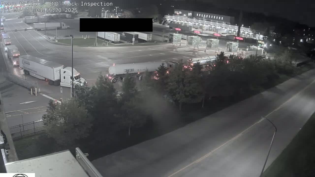At night, several semi-trucks are at a border crossing inspection area, with one semi-truck and tanker positioned for inspection.