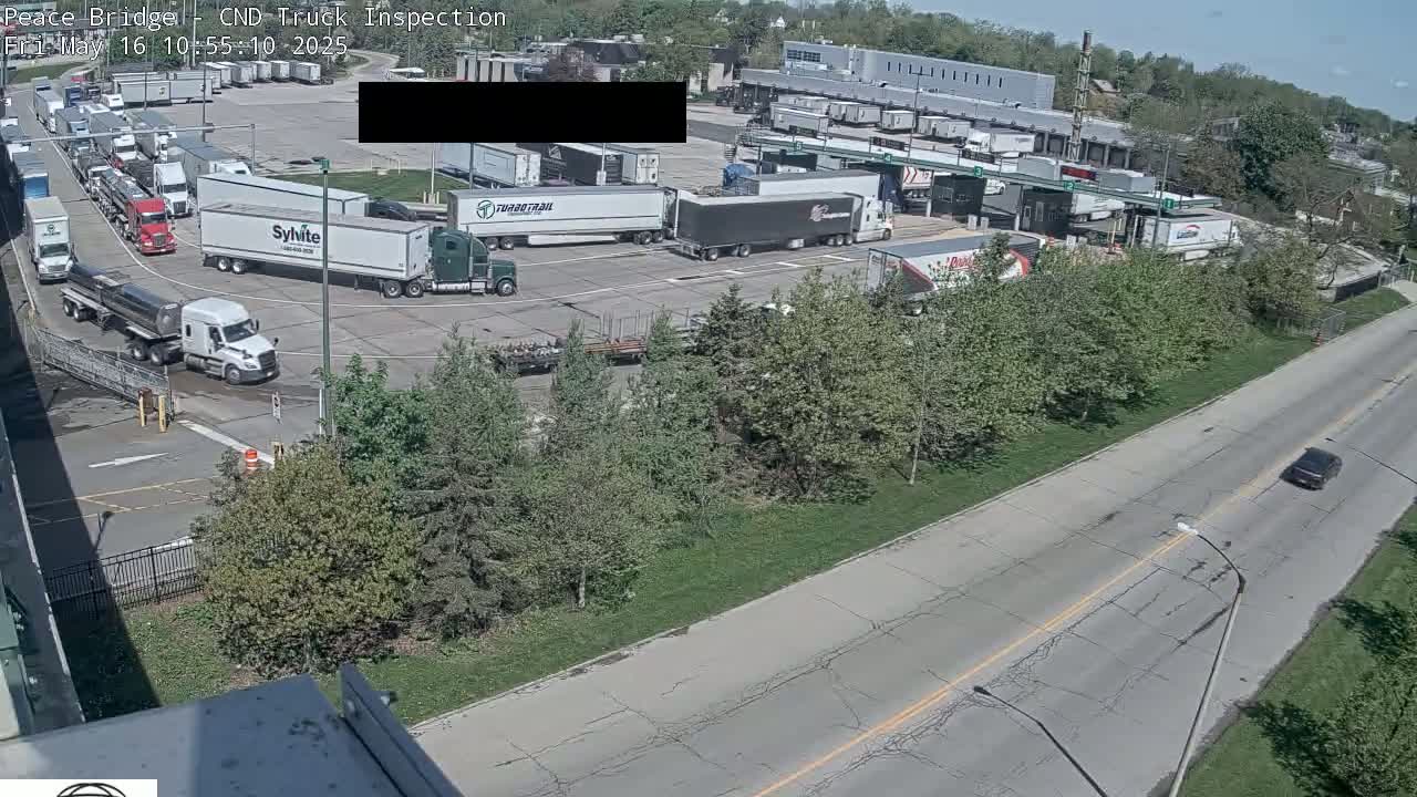 A truck inspection station with numerous semi-trucks and trailers parked and in line, on a sunny day, viewed from an elevated vantage point overlooking a road with a single car.
