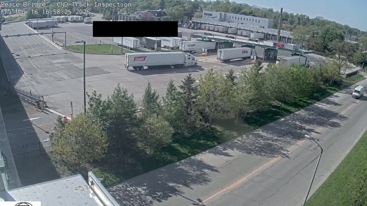 A high-angle view shows a truck inspection area with several semi-trucks parked in a sunny, outdoor lot next to a road lined with trees.