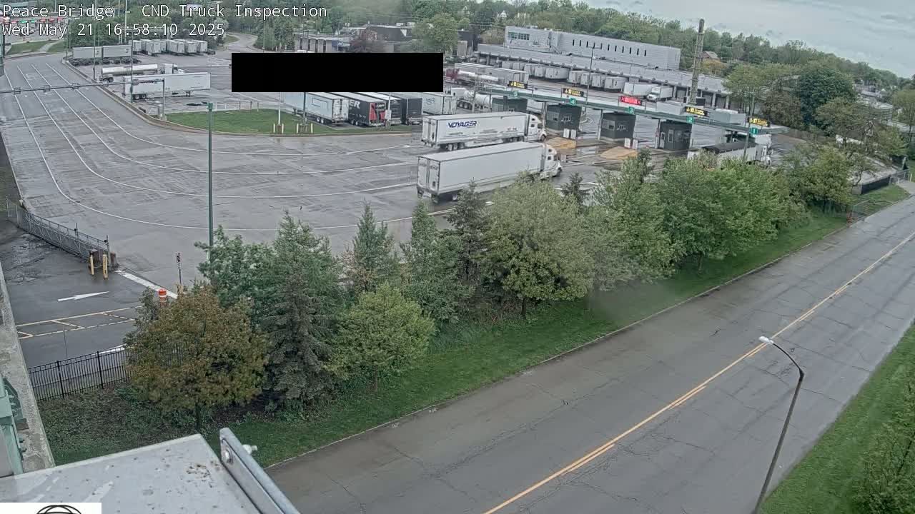A wet, paved area with several semi-trucks parked near a border crossing, surrounded by green trees and shrubs under an overcast sky.