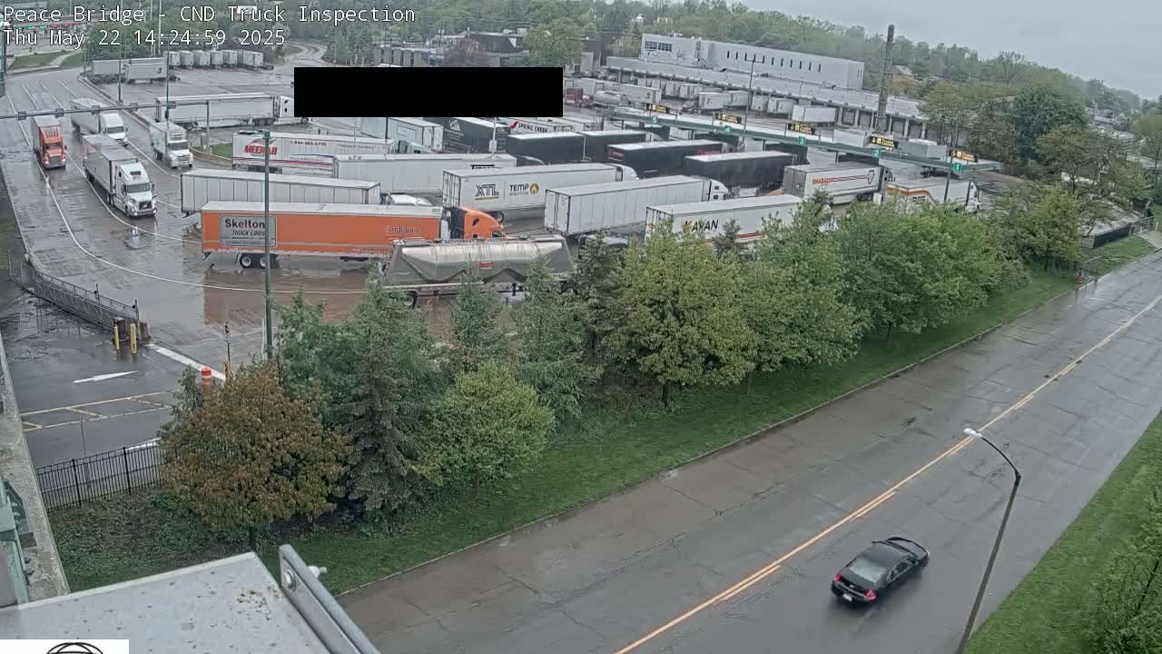 A wet, paved area with numerous semi-trailer trucks parked and a single car driving on a road next to a grassy area with trees.