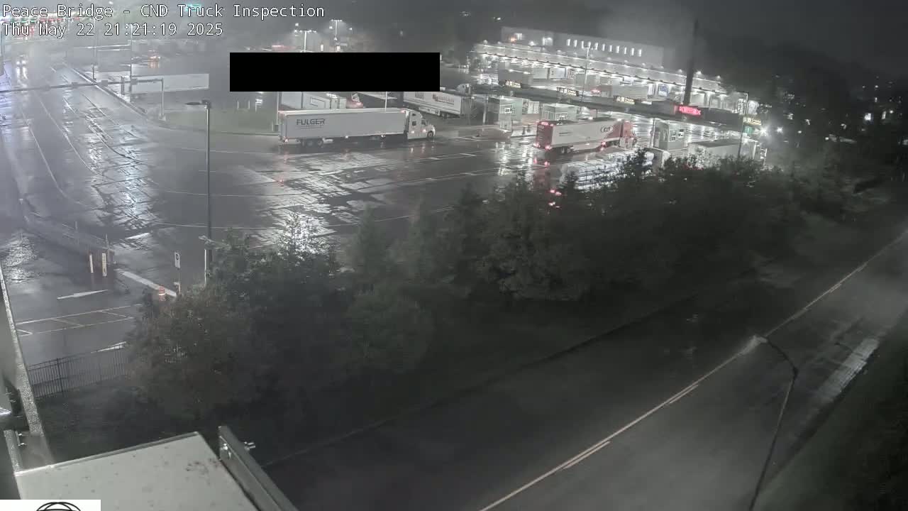A nighttime, rain-slicked truck inspection area shows several semi-trucks parked and moving in the lot, with surrounding roadways and vegetation visible.