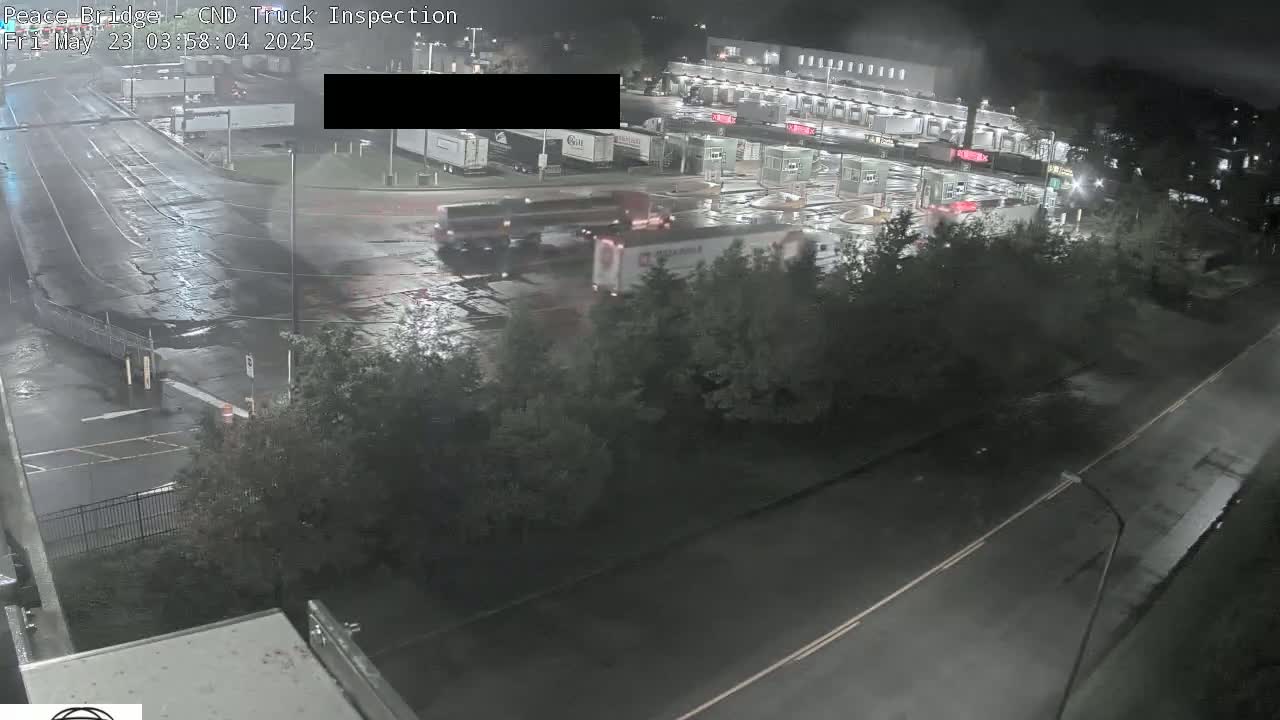 Several semi-trucks are maneuvering in a wet, nighttime roadway near a border crossing facility.