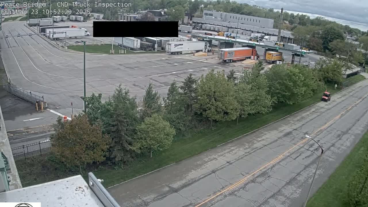 An aerial view shows a truck inspection station with several semi-trucks parked and a road with some light traffic under an overcast sky.