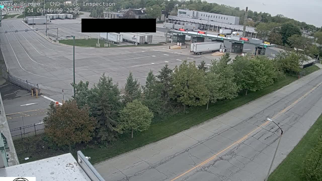 An aerial view shows a truck inspection station with several semi-trucks, a roadway with a dividing line, and a row of trees under an overcast sky.