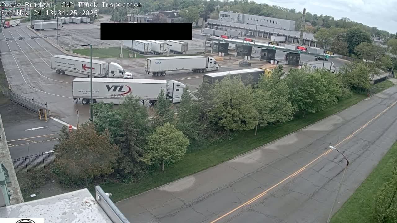 Several semi-trucks are parked and moving in a large paved area next to a road lined with trees under an overcast sky.