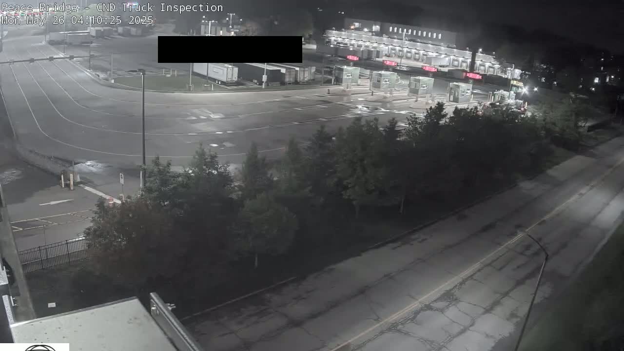 A nighttime, overhead view shows a truck inspection station with several semi-trucks parked nearby and a roadway with minimal traffic.