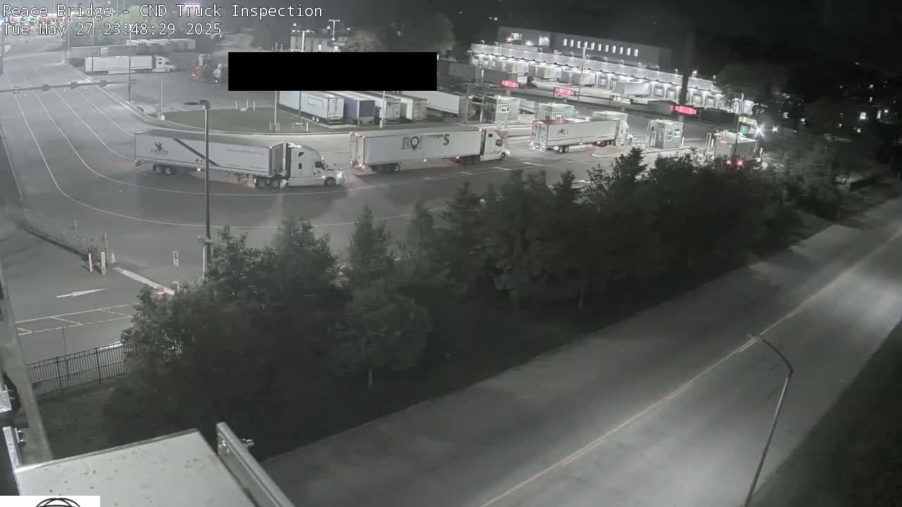 Several semi-trucks are parked and moving at night in a large, well-lit outdoor area, surrounded by trees and a roadway.
