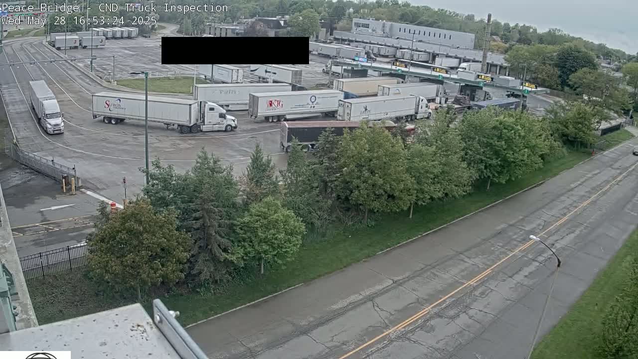 An overhead view shows a truck inspection area with numerous semi-trailer trucks parked and maneuvering on a wet, overcast day.