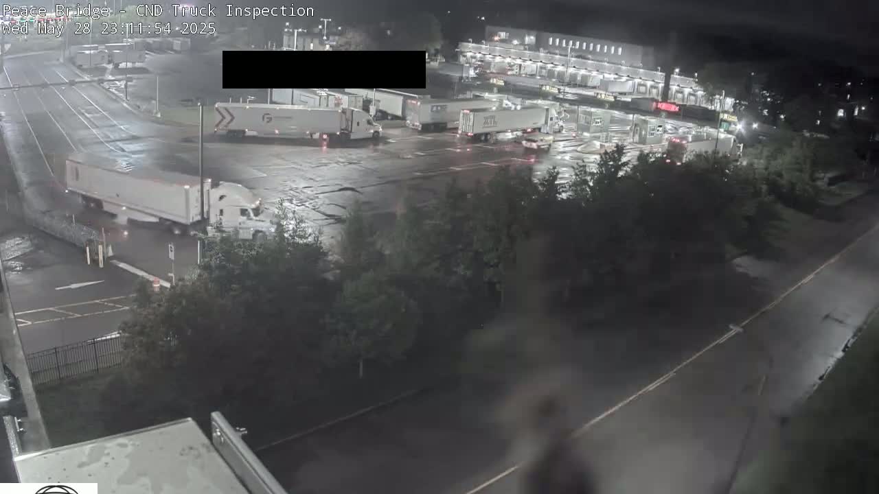 Several semi-trailer trucks are parked and driving in a large, wet asphalt lot at night.